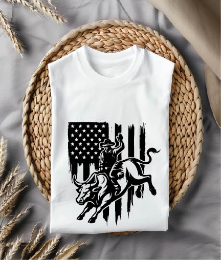 White t-shirt with a graphic design of a cowboy on a bull and an American flag, placed on a woven mat.