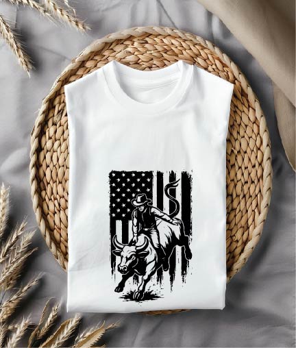 White t-shirt with a graphic design of a cowboy on a bull and an American flag, placed on a woven mat.