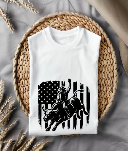 White t-shirt with a graphic design of a person riding a bull on an American flag, placed on a woven mat.