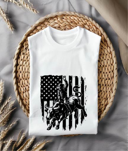 White t-shirt with bull riding design on an American flag background, placed on a woven mat.