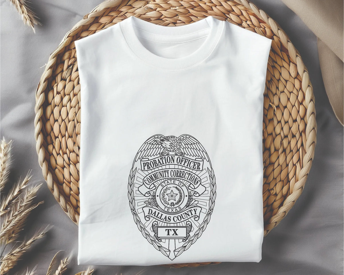 A white short-sleeve T-shirt folded flat on a woven straw mat, with a black line-art badge graphic on the front reading 'Probation Officer' and 'Dallas County TX.'.