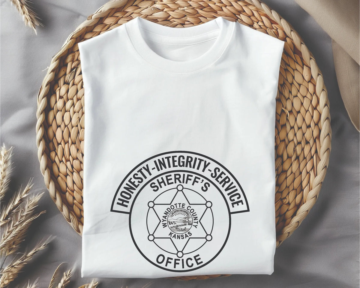 A folded white crewneck t-shirt with a black circular graphic reading 'HONESTY-INTEGRITY-SERVICE SHERIFF'S WANDITE COUNTY KANSAS OFFICE', displayed on a woven straw mat over a gray fabric background with dried wheat stalks.