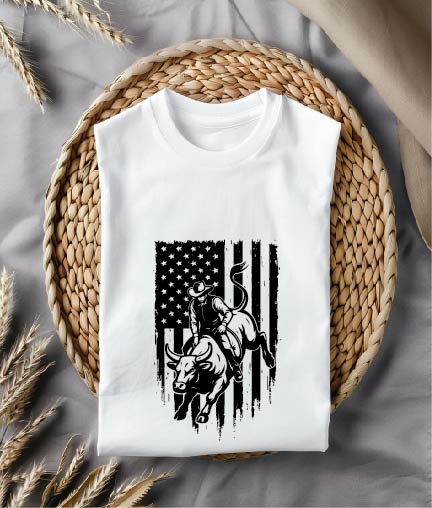 White t-shirt with a graphic design of a cowboy on a bull in front of an American flag, placed on a woven mat.