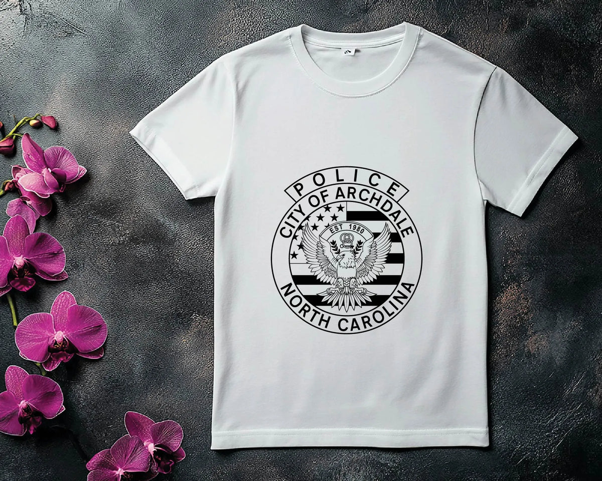 A white short-sleeve crewneck t-shirt featuring a black circular graphic of the City of Archdale Police seal from North Carolina, styled flat-lay on a dark textured surface with pink orchids on the left.