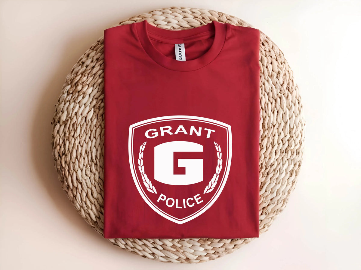 A folded red t-shirt with a white 'GRANT POLICE' shield graphic on the front, displayed on a round woven straw mat against a light beige background.
