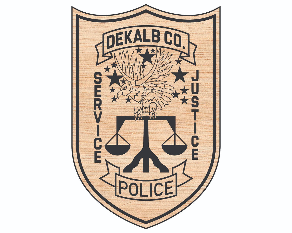 A shield-shaped wooden plaque or sign featuring a black engraved emblem with an eagle, stars, scales of justice, and the text 'DEKALB CO. SERVICE JUSTICE POLICE' on a light natural wood background.