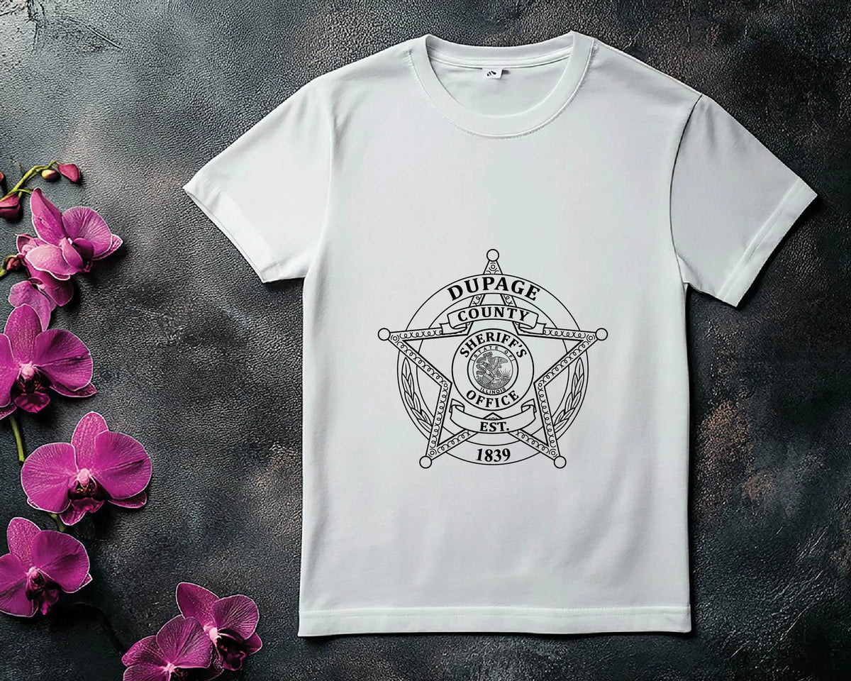 A white short-sleeve crewneck t-shirt featuring a black and gray Dupage County Sheriff's Office graphic on the front, styled flat-lay on a dark textured surface with purple orchids on the left.